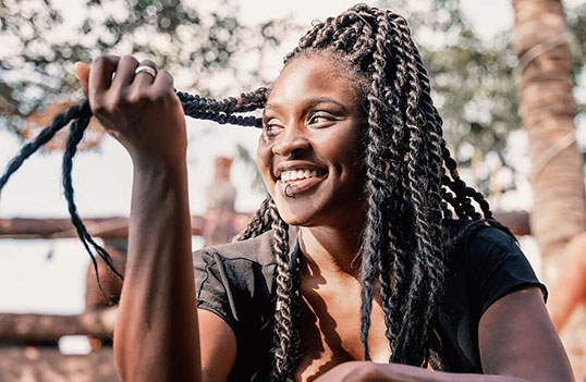 A person with long, braided hair holds a section of their braid while seated outdoors, surrounded by trees and soft sunlight.