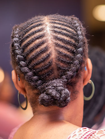 Close-up of a woman's braided hairstyle featuring intricate cornrows leading to a bun at the nape of her neck, showcasing texture and detail.