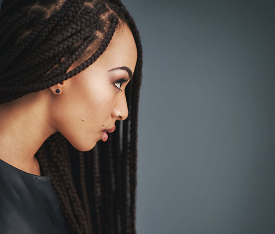 A person with long, braided hair turned slightly to the side, showcasing a simple black shirt against a soft, muted background.