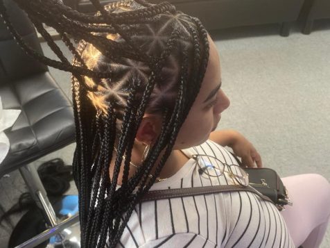 A person with long braided hair styled upwards sits in a salon, wearing a striped shirt and holding a phone. Bright lighting highlights the hair.