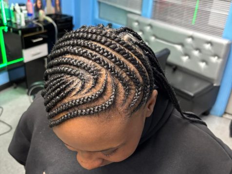 A close-up view of intricately styled cornrow braids forming a spiral pattern on a person's scalp, in a vibrant barbershop setting.
