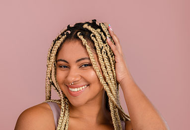 A person with long braided hair poses against a soft pink background, gently touching their head with one hand.