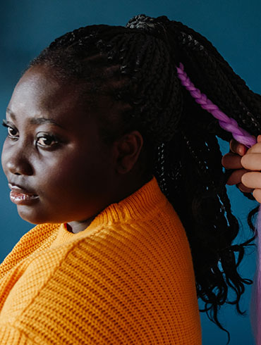 A person with braided hair styled in a high ponytail, wearing a bright orange sweater against a teal background.